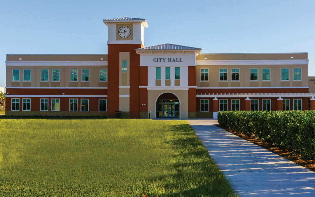 A modern, two-story building with a clock tower labeled "City Hall" stands proudly in Palm Coast. The structure has large windows and is surrounded by a lush green lawn. A paved path leads from the lawn to the entrance, and the sky is clear and blue, making it a picturesque scene in Flagler County.