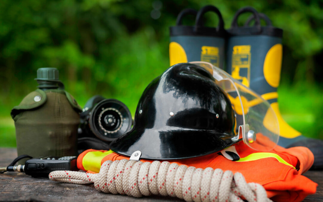 A collection of firefighting gear is shown, including a black helmet with a clear visor, a pair of yellow and black boots, a green canteen, gloves, a rope, and an orange outfit with reflective strips. All are laid out on a wooden surface with greenery in the background in Palm Coast.