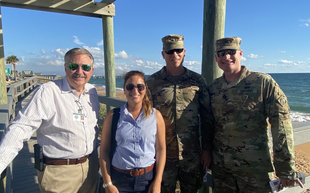 Four people are standing on a boardwalk by the beach in Palm Coast, with the ocean in the background. Two individuals in the center are wearing military uniforms, while the person on the left is in a white shirt and khakis, and the person on the right is in a blue blouse and sunglasses.