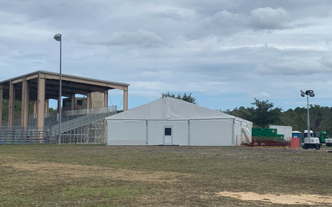 A large white tent is set up on a grassy field near a wooden stadium structure with bleachers in Palm Coast. Some trees, a utility vehicle, and several portable units are visible in the background under a cloudy sky. This scene recently made headlines in Flagler County news.