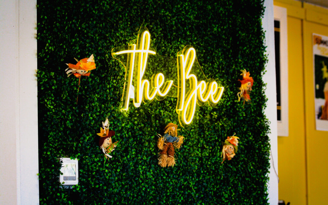 A wall covered in green artificial foliage with a bright yellow neon sign that reads "The Bee" evokes a charming Palm Coast vibe. Small scarecrow decorations are attached to the foliage, and a light switch is visible on the lower left side. In the background, there is a partially visible yellow wall.