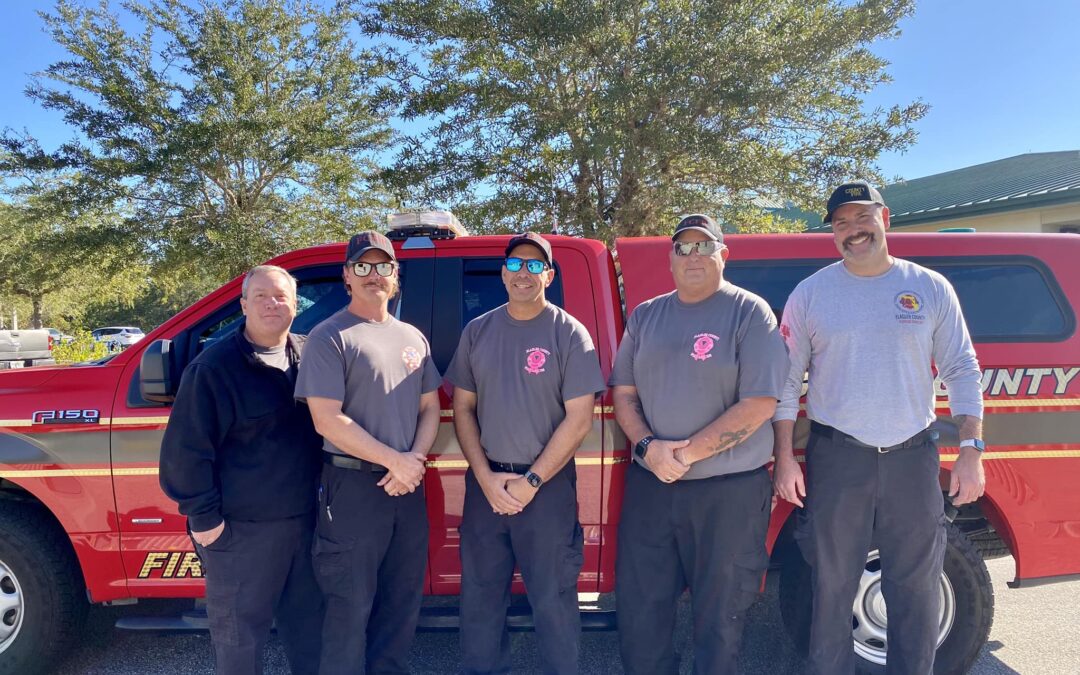 A group of five firefighters from Palm Coast stands in front of a red fire truck. Four wear gray t-shirts, black pants, and caps, while one wears a black jacket. They are outdoors on a sunny day in Flagler County, with trees and a building in the background.