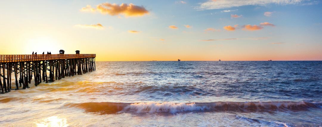 A long wooden pier extends over the ocean at sunset in Palm Coast, with a few people walking on it. The sky is painted in warm shades of orange, pink, and blue, with scattered clouds. Gentle waves lap at the shore in Flagler County, reflecting the colorful sky.