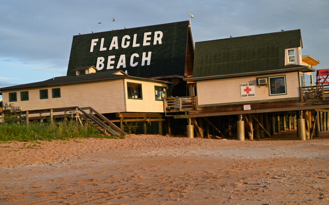 A beach building on stilts with a large sign reading "Flagler Beach" in bold white letters. There's a lifeguard station with a red cross sign on the right side of the building. The sandy beach and an overcast sky create a calm coastal scene in Flagler County, near Palm Coast.