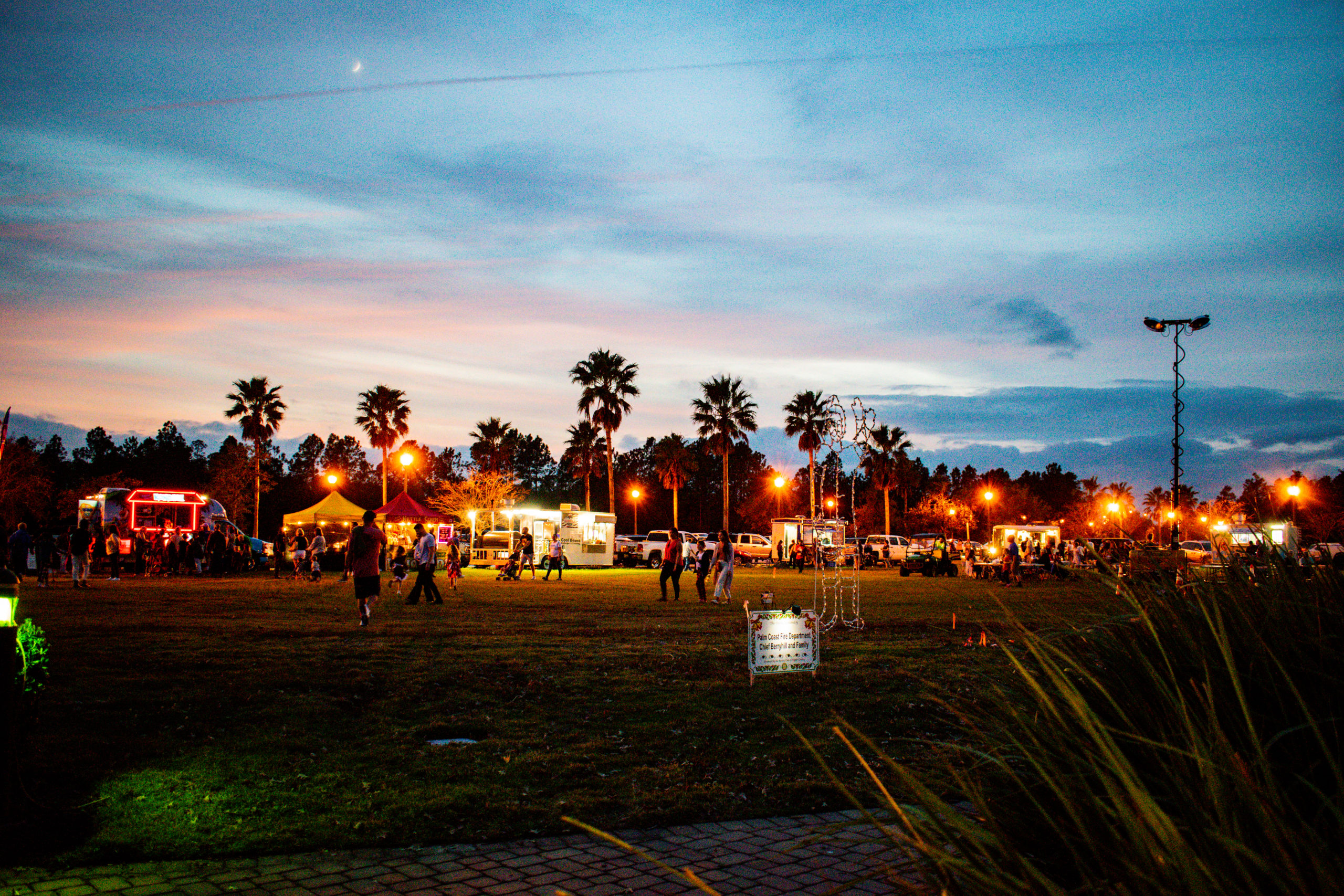 An outdoor evening event in Palm Coast with people gathered on a grassy field surrounded by palm trees. Food trucks, illuminated with lights, dot the area as the sky is painted with hues of blue and pink at sunset. A few tents and a tall light pole are also visible, adding to the serene ambiance in Flagler County.