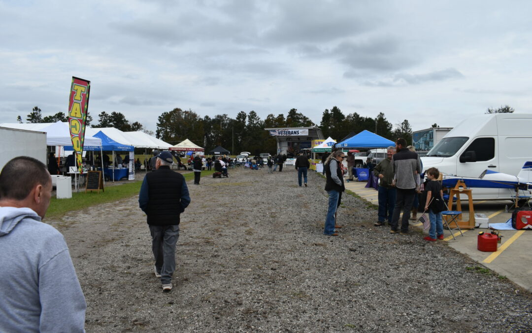 Outdoor market scene with people walking along a gravel pathway. Various vendor stalls are set up, selling items like food and crafts. A "Tacos" sign is visible, and there are tents and vans around. Trees and cloudy skies form the backdrop. It's a bustling event in Palm Coast, Flagler County.