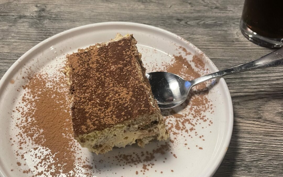 A slice of tiramisu topped with cocoa powder is on a white plate with a spoon resting next to it. The plate is on a wooden table in Palm Coast, with a glass of dark beverage to the right and a small plate with whipped cream and a pastry in the background.