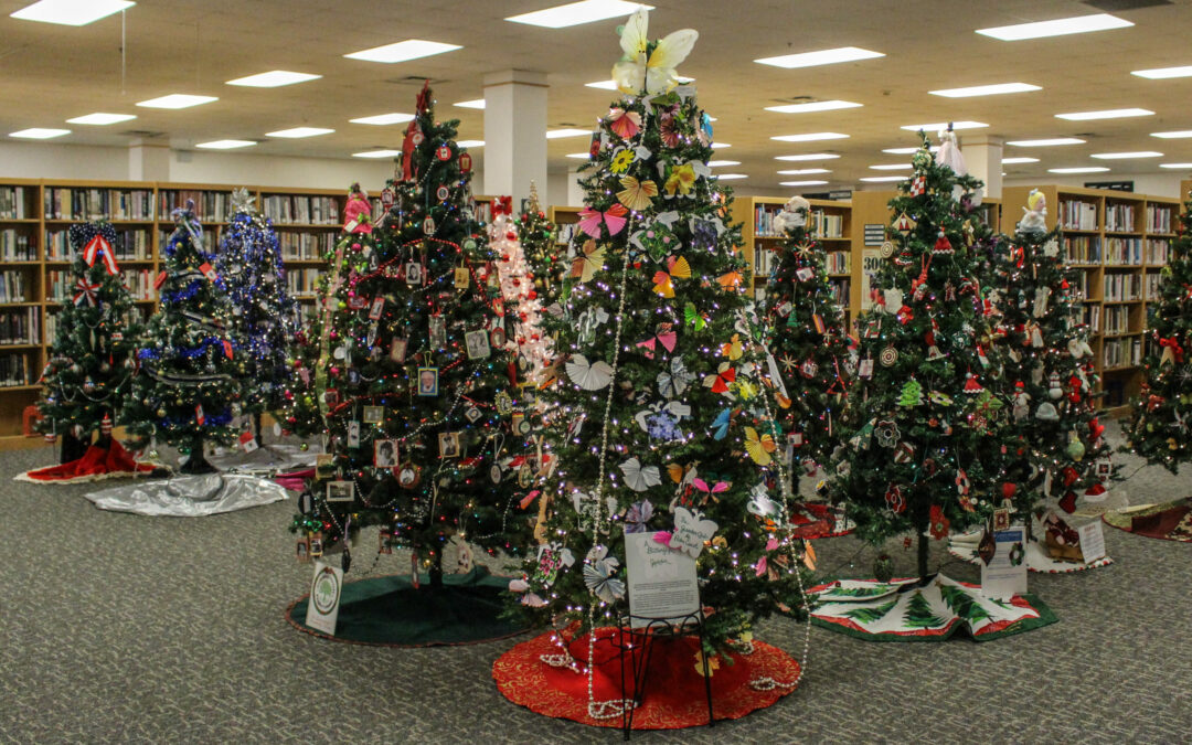 A room in a Palm Coast library is filled with various decorated Christmas trees, each adorned with ornaments, bows, and lights. Bookshelves line the back walls. The carpeted floor and well-lit area create a warm holiday atmosphere perfect for Flagler County residents.