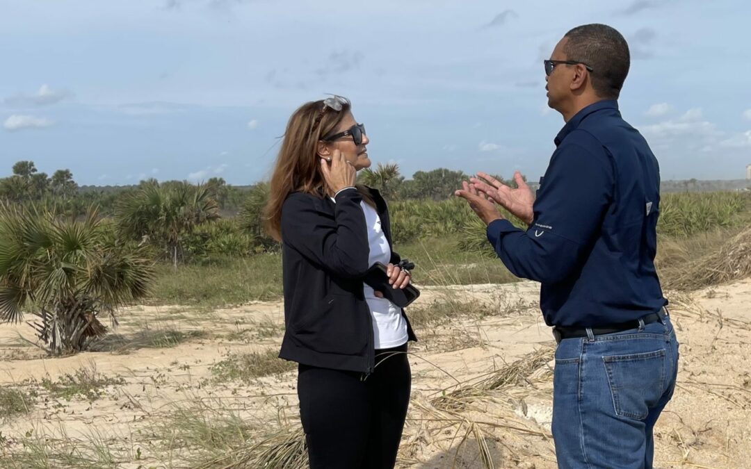 Two people are standing on a sandy terrain in Flagler County, engaged in conversation. The woman on the left has long hair, sunglasses, and is dressed in a black jacket, black pants, and red boots. The man on the right is in jeans, a blue shirt, and sunglasses. The background is a clear sky and sparse vegetation.