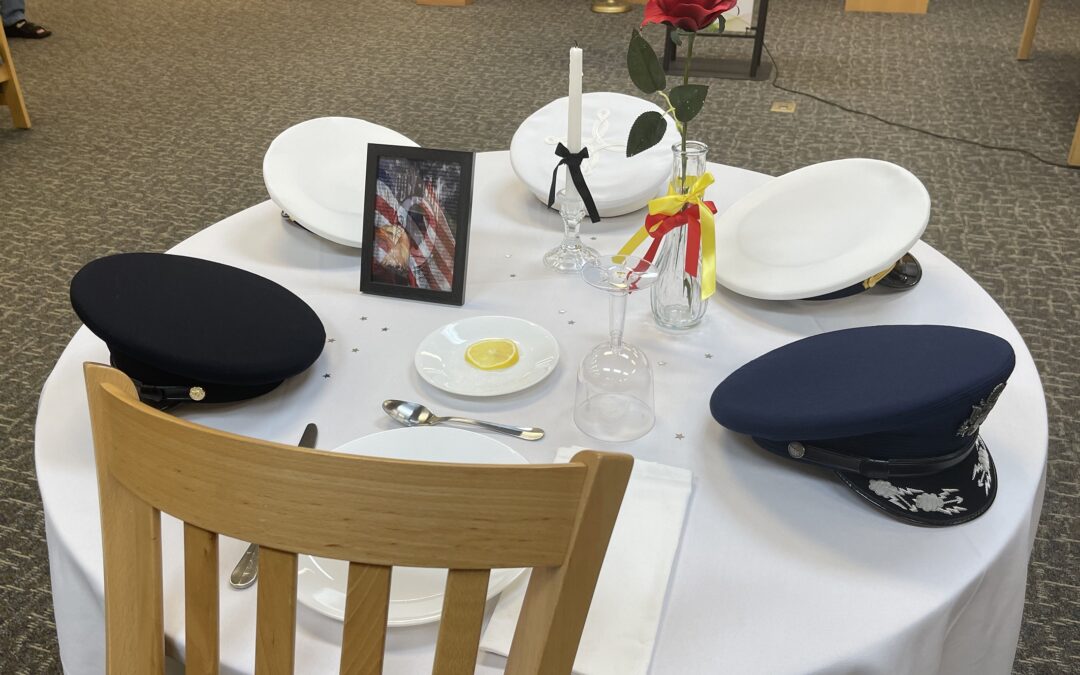 In Palm Coast, a round table is set with four military hats, a framed photo of an American flag, a single lit candle, a red rose in a vase, and an empty plate with a lemon slice and salt. The tribute honors missing servicemembers and POWs in Flagler County.