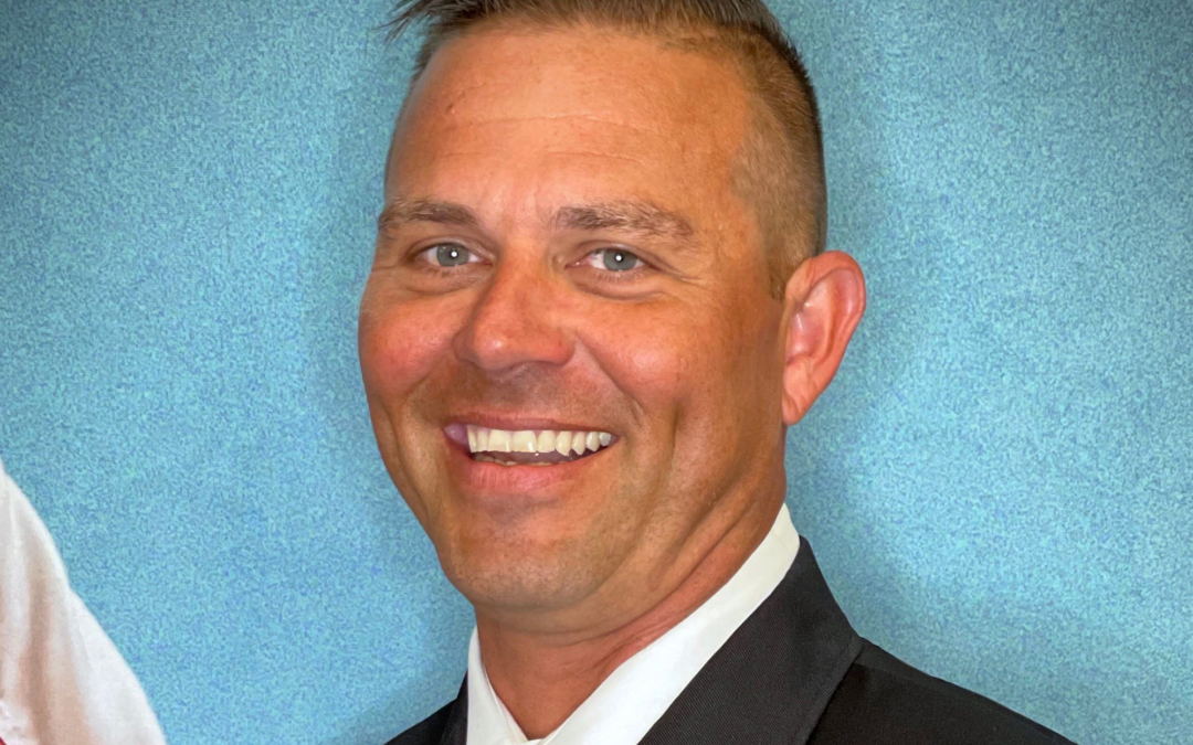 A man in a dark formal uniform is smiling, with his head turned slightly to the right. He has short hair and is wearing two red pins on his lapel. The background is a gradient blue with a hint of the American flag visible on the left side, representing Palm Coast news in Flagler County.