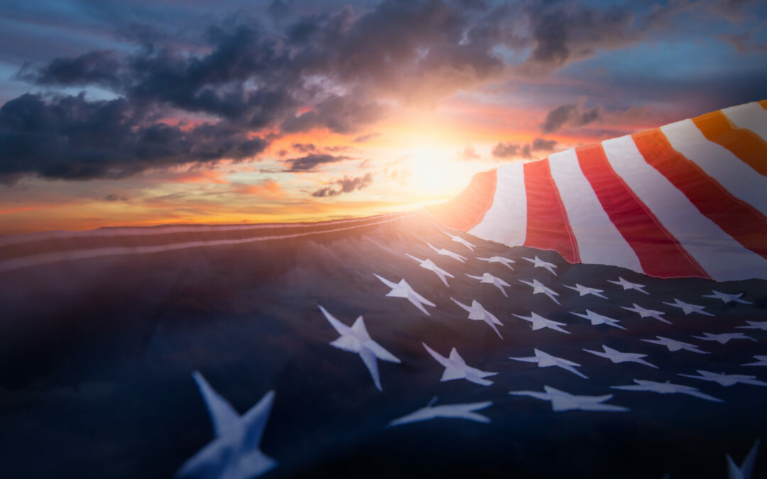 An American flag waves prominently in the foreground, partially obscuring a dramatic Palm Coast sunset with vibrant orange and purple hues. Dark clouds are scattered across the Flagler County sky, enhancing the striking contrast of colors.