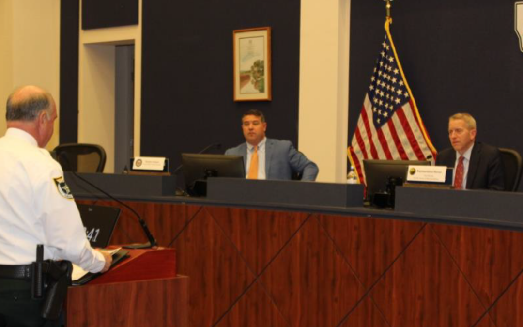 A sheriff in uniform stands at a podium addressing two seated officials in a Flagler County government meeting room. An American flag and Palm Coast logo are visible in the background. The officials are seated at a long desk with nameplates, lending a formal tone to the local news update.