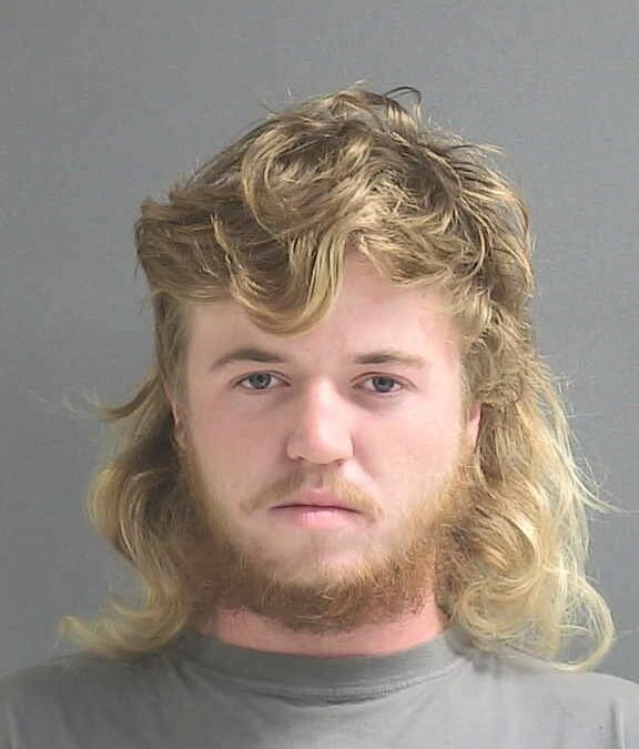 A young man with long, curly, light brown hair and a beard looks directly at the camera with a neutral expression. He is wearing a grey shirt and is photographed against a plain, grey background. The photo appears to be for a Palm Coast news feature in Flagler County.