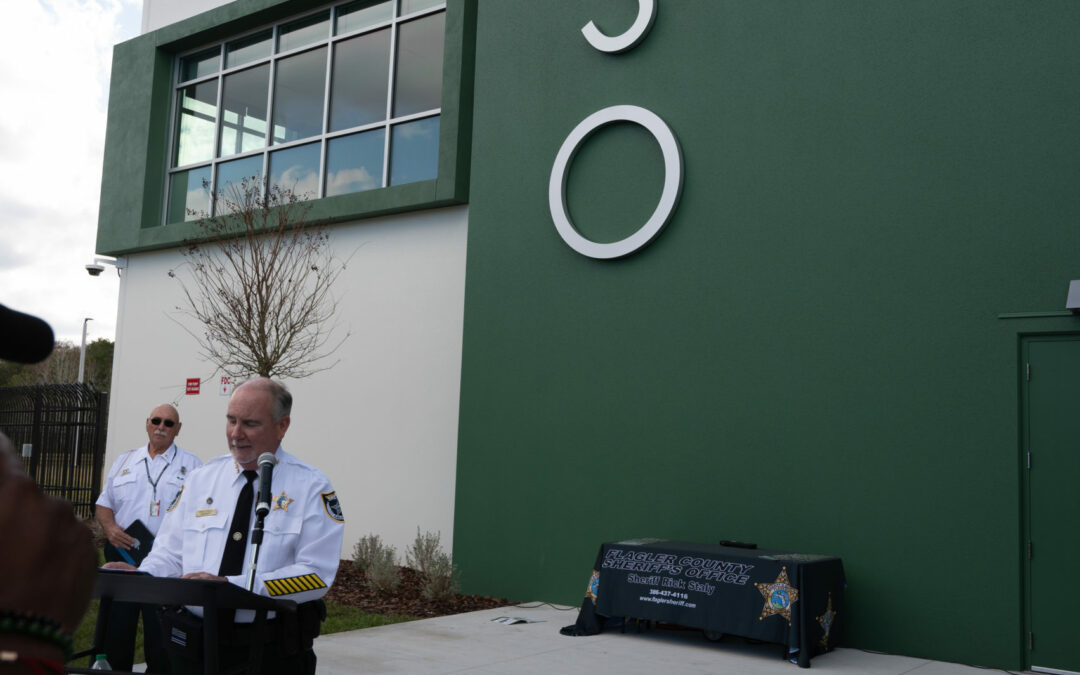 A law enforcement officer in uniform stands at a podium giving a speech outdoors in front of a building with large white letters reading "FCSO." A table with a dark blue logo cover is visible in the background. Another officer stands near a door to the right. This scene unfolds in Palm Coast, Flagler County.