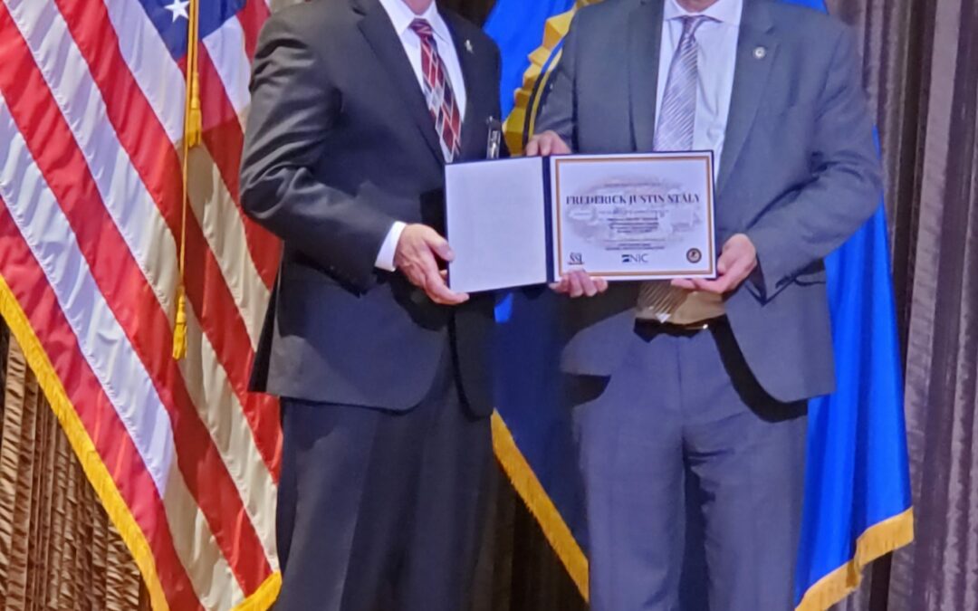 Two men in suits stand on a stage. One holds an open certificate, and the other smiles beside him. Behind them, American and other flags are displayed. The formal setting suggests a ceremony or award presentation, potentially in Flagler County or Palm Coast.