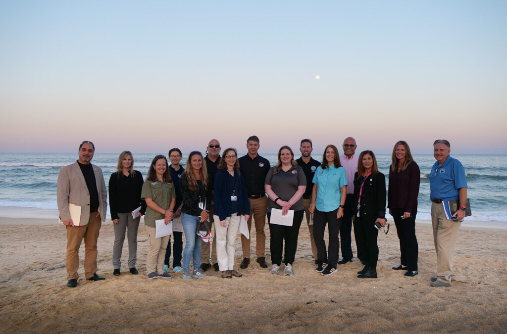 A group of fifteen people posing on a sandy beach in Palm Coast with the ocean and a colorful sunset sky in the background. They are dressed in a mix of casual and business casual attire, some holding papers. The mood appears friendly and relaxed, capturing a quintessential Flagler County evening.