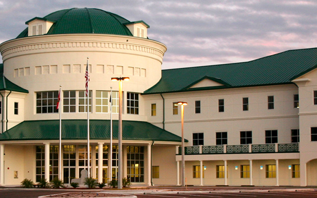 A large, multi-story building with a white facade and a green domed roof is pictured in the heart of Palm Coast, Flagler County. The structure features several windows, a main entrance with multiple glass doors, and an array of flagpoles out front. The sky above is cloudy, casting a soft light.