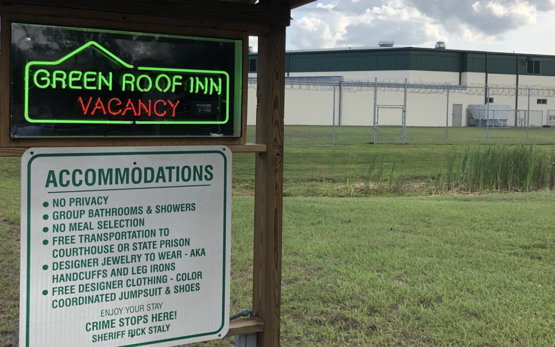 A neon sign reads "Green Roof Inn Vacancy" beside another sign listing sarcastic "accommodations" like no privacy, group bathrooms and showers, free transportation to court or prison, and enforced clothing, clearly referring to a jail in Flagler County. The Palm Coast institution's building and fencing are visible.