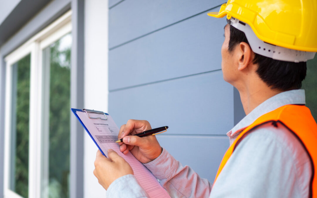 A construction worker in a yellow hard hat and orange safety vest is facing a grey-blue building in Palm Coast, Flagler County, holding a clipboard and writing on a piece of paper. The worker appears to be inspecting or documenting something related to the building's exterior.