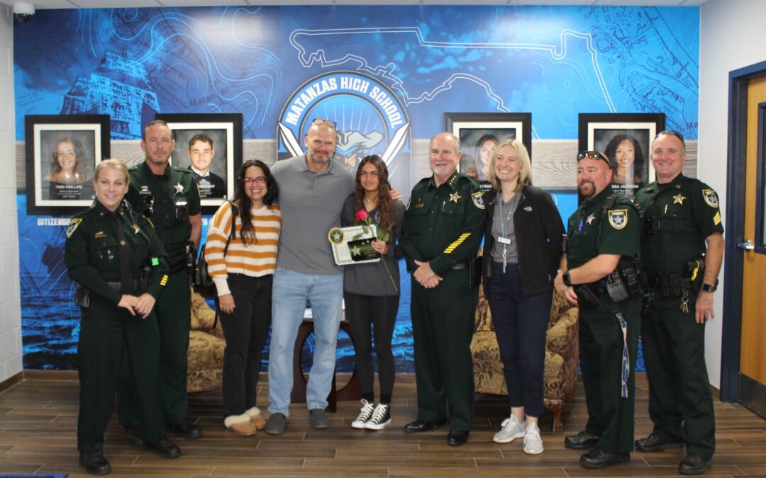 A group of people, including several uniformed officers and other adults, pose together in a room with blue walls and framed photographs. One person holds a certificate or plaque. The backdrop features "Manatees High School" in bold. Smiles and relaxed poses indicate a celebratory occasion in Palm Coast, Flagler County.