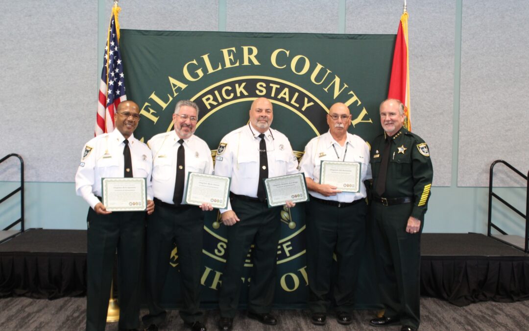 Five uniformed officers stand in front of a Flagler County Sheriff's Office banner in Palm Coast. Four hold certificates, while the fifth is empty-handed. They stand in front of an American flag and a red flag with a seal, all smiling for the photograph. This news captures their proud moment.