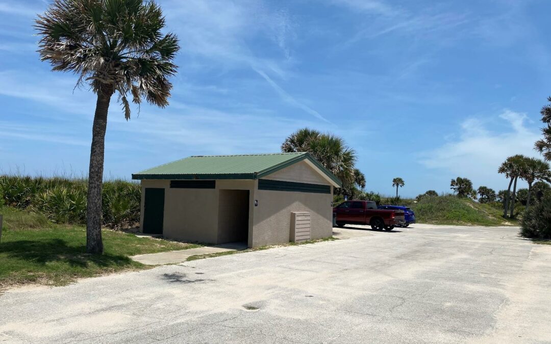 A small building with a green roof stands next to a palm tree in a parking lot under a clear blue sky in Palm Coast. Two vehicles, a red SUV and a blue truck, are parked near the building. In the background are more palm trees and grassy dunes, typical of Flagler County's scenic views.