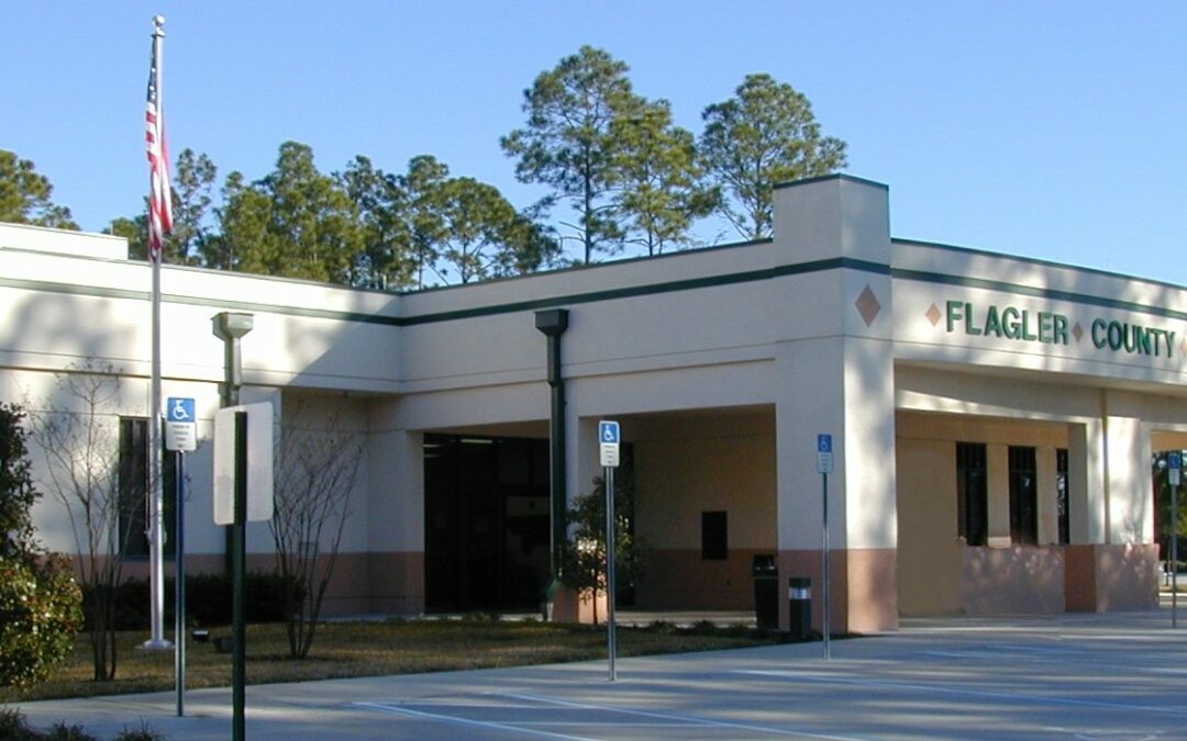 A single-story building labeled "Flagler County Public Library" with an American flag in front. The structure features a covered entrance, beige and white walls with green accents, and is surrounded by a landscaped area including trees and shrubs, making it a focal point in the Palm Coast community.