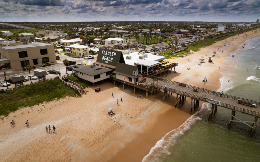 A coastal town in Flagler County features a sandy beach with a pier extending over the ocean. Beachgoers relax under umbrellas and swim in the water. Buildings, including one labeled "F-L-A-G-L-E-R BEACH," are situated near the shore under a partly cloudy sky.