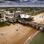 A coastal town in Flagler County features a sandy beach with a pier extending over the ocean. Beachgoers relax under umbrellas and swim in the water. Buildings, including one labeled "F-L-A-G-L-E-R BEACH," are situated near the shore under a partly cloudy sky.