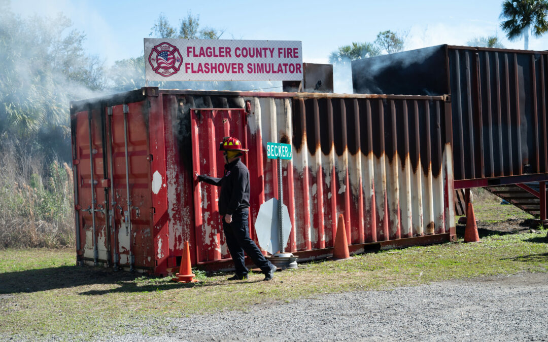 A firefighter wearing a helmet and gear stands at the entrance of a smoking container labeled "Flagler County Fire Flashover Simulator." The container, showing signs of heat exposure, is part of a Palm Coast training site, with safety cones placed nearby.