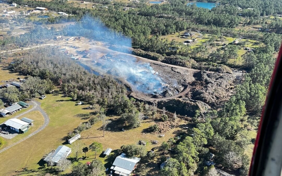 Aerial view of a semi-rural landscape in Palm Coast, Flagler County, with homes, trees, and open fields. Smoke rises from a burning area amid the greenery, indicating a fire. Surrounding the area are various structures and patches of forests, with clear skies overhead. News crews are reporting live.