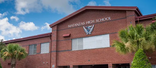 A two-story red brick building with "Matanzas High School" written on a sign near the top. The building, located in Palm Coast, features a large metal emblem and has several windows with shutters. The front is landscaped with palm trees and bushes, and the sky above is partly cloudy—a true Flagler County gem.