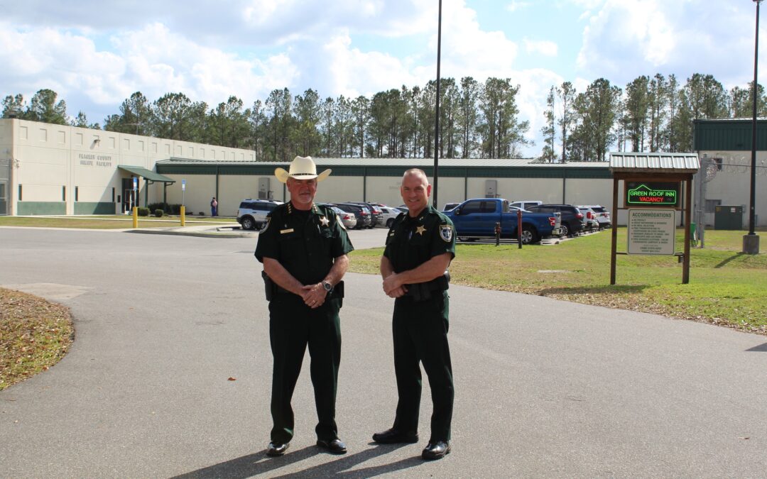 Two uniformed law enforcement officers, one wearing a cowboy hat, stand together in a parking lot outside a large building with a sign reading "County Jail." Cars are parked in front of the building in Flagler County, and trees are visible under a partly cloudy sky.