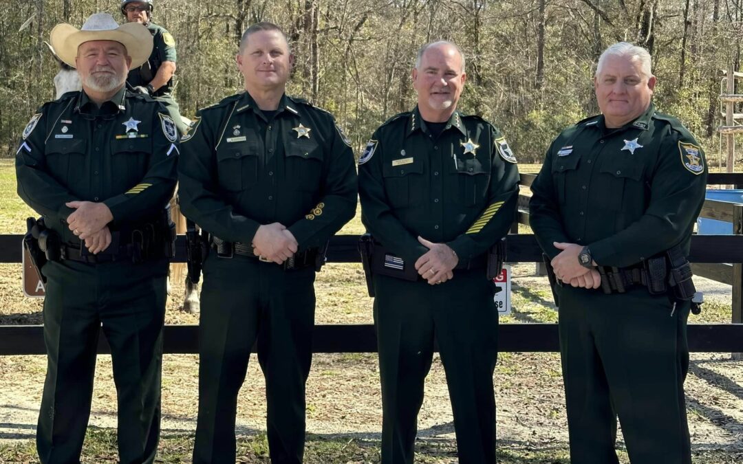 Four male officers in green uniforms with star badges stand in a line on a dirt path, with the black wooden fence and trees of Flagler County in the background. One wears a cowboy hat while another sits on horseback. This scene is set in Palm Coast and looks like something right out of the news.