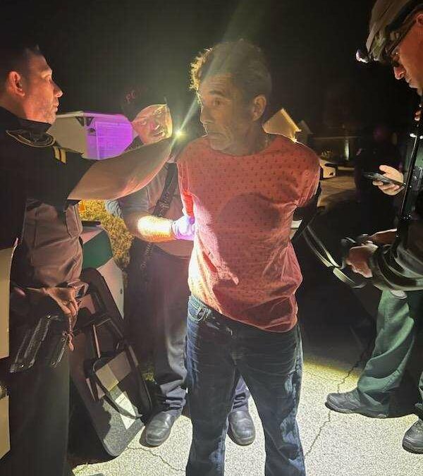 A man in a red shirt stands with his hands behind his back, detained by officers at night. Two uniformed officers are speaking to him while another in a helmet stands beside them. The area appears to be illuminated by car headlights, casting dramatic lighting over the scene in Flagler County.