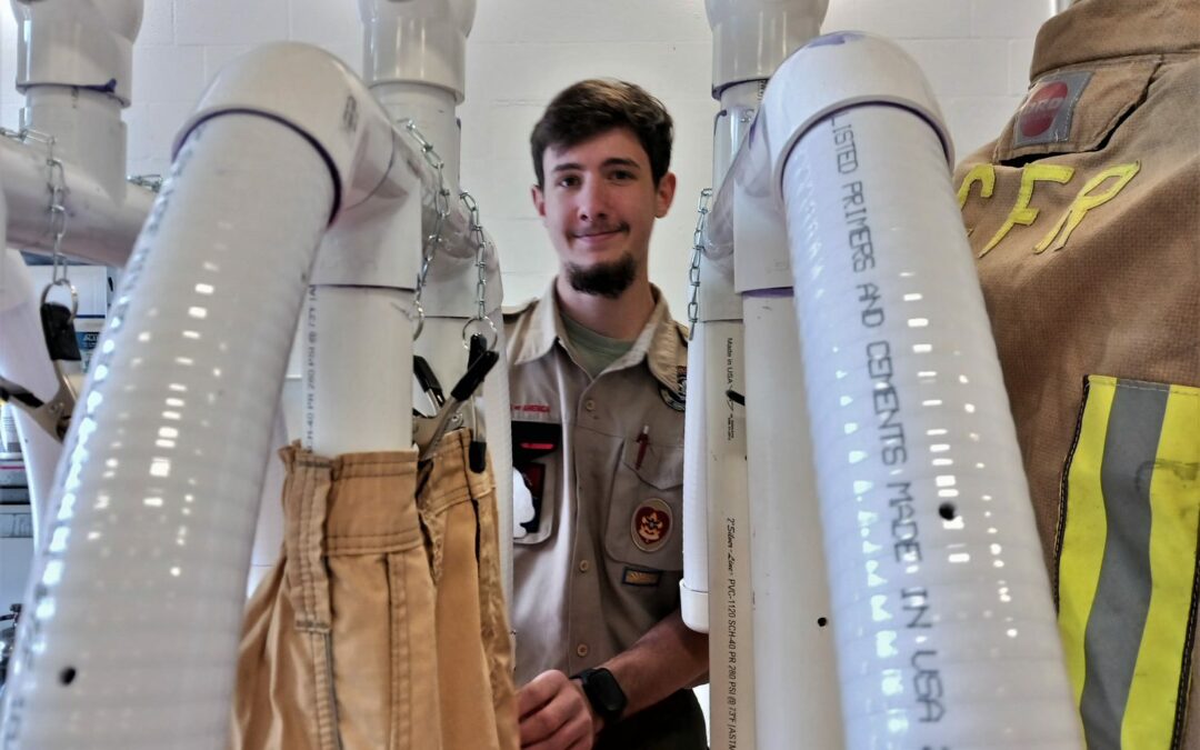 A man stands between drying racks holding beige garments, with industrial drying machines in the foreground. He wears a uniform and is surrounded by equipment, including firefighter gear with "CFR" visible on a jacket hanging to the right, in this scene from Palm Coast, Flagler County.