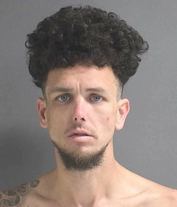 A man with curly hair and a goatee is standing against a plain gray background. He has blue eyes and a neutral expression. Shirtless, revealing tattoos on his left shoulder, he could easily be a notable figure from Flagler County or Palm Coast, making headlines in local news.