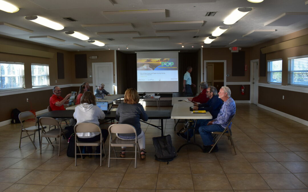 A group of people are seated around tables arranged in a U-shape in a brightly lit room with tiled floors in Flagler County. They are facing a presentation screen showing a slide. Laptops and papers are on the tables, and some individuals are taking notes or looking at the screen.