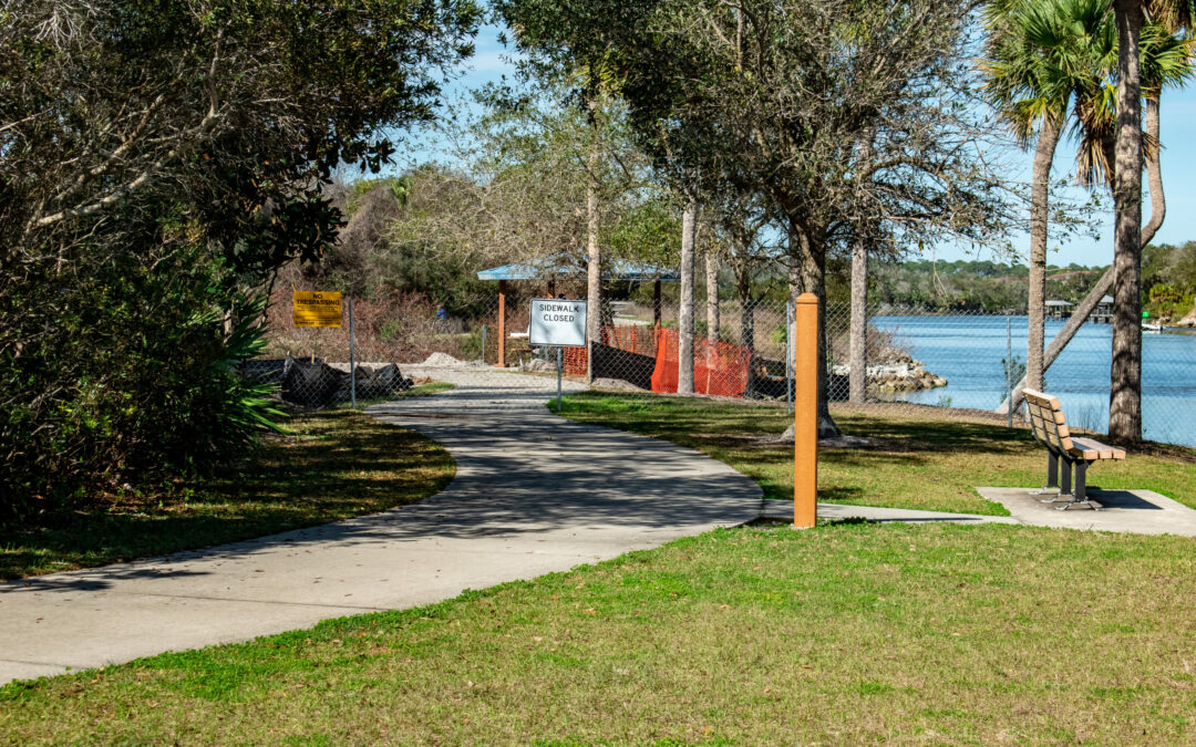 A paved pathway winds through a park in Flagler County, dotted with trees and a bench on the right side. The path leads to a fenced area near the water, where a sign reads "DOCKING CLOSED." Surrounded by lush greenery, this serene Palm Coast spot offers a scenic view of the water in the background.