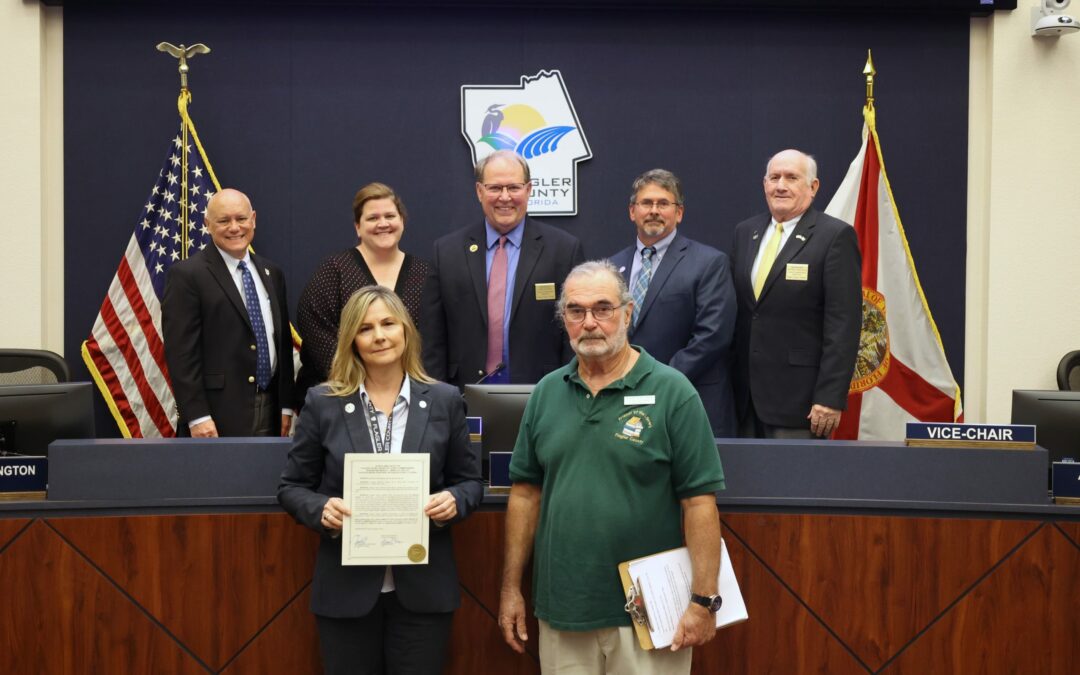 A group of seven people posing indoors in an official setting in Palm Coast. Four men and one woman stand behind a woman and a man who are holding a document. Flags are visible in the background against a backdrop with a logo, highlighting the importance of this moment for Flagler County.