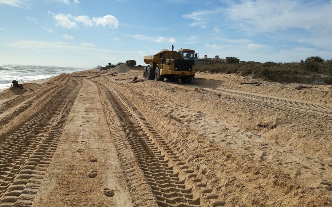 A large yellow bulldozer is parked on a sandy beach in Palm Coast, with deep tread marks leading up to it. The sky above is partly cloudy with some areas of blue. Vegetation lines the right side of the beach, and the ocean is visible on the left—just another day in Flagler County news.