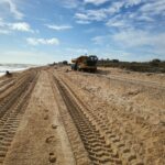 A large yellow bulldozer is parked on a sandy beach in Palm Coast, with deep tread marks leading up to it. The sky above is partly cloudy with some areas of blue. Vegetation lines the right side of the beach, and the ocean is visible on the left—just another day in Flagler County news.