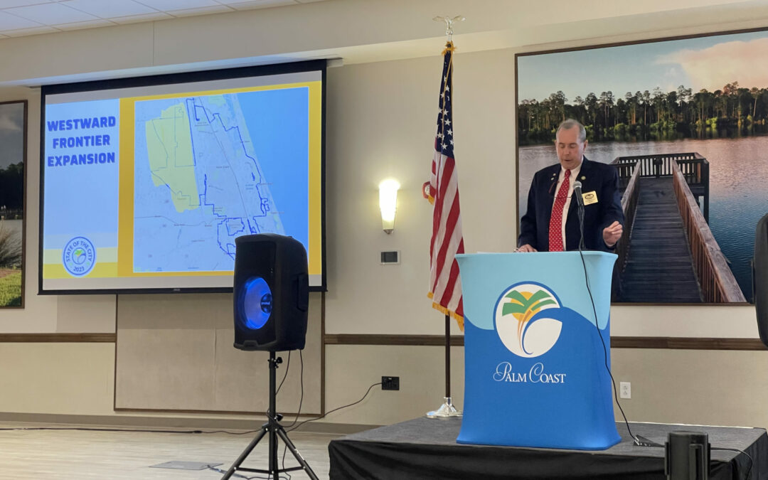 A man in a suit stands at a podium with a microphone, speaking in a conference room. He is gesturing towards a projection screen displaying a map labeled "Westward Frontier Expansion." An American flag is placed beside the podium, which displays the Palm Coast logo, highlighting an important moment for Flagler County.