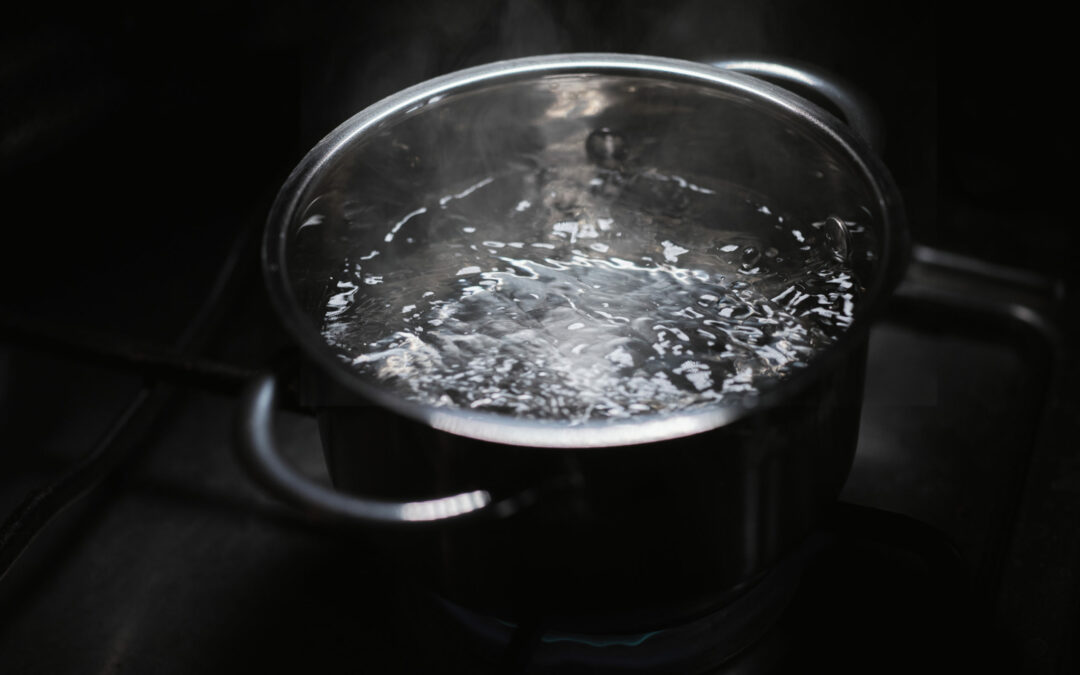 A silver pot filled with boiling water is placed on a stove, bubbling vigorously as steam rises. The dark background emphasizes the bright, shiny surface of the pot and the movement of the boiling water. This scene might evoke a quiet evening in Palm Coast, Flagler County.