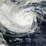 A satellite image of a massive, swirling hurricane over the ocean captures the eye of the storm at its center, surrounded by thick white clouds spiraling outward. With counterclockwise rotation, the dark and turbulent ocean below appears ominous. This significant weather event is now impacting Flagler County and Palm Coast.