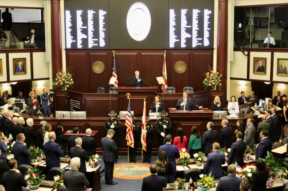 A legislative chamber filled with delegates in formal attire stands as an official speaks at a podium beneath a large electronic display. Two flags are placed near the center. Floral arrangements decorate the space, and various individuals observe from balconies above, capturing attention in Flagler County news outlets.