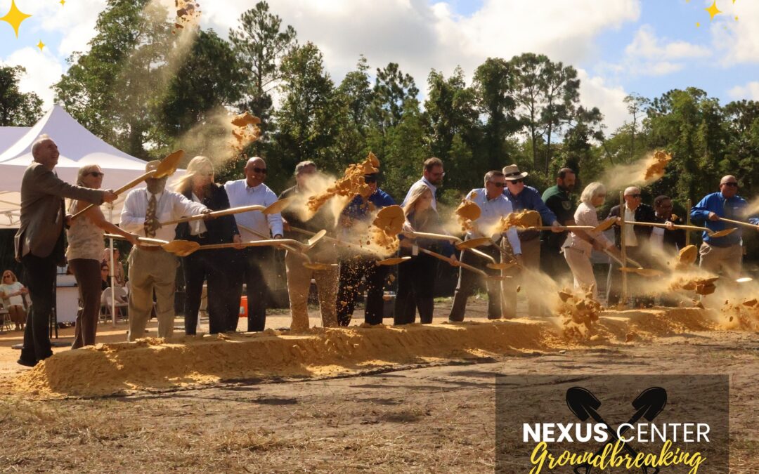 A group of people stand in a row with shovels, tossing dirt during a groundbreaking ceremony outdoors in Flagler County A sign at the bottom right reads "Nexus Center Groundbreaking" with the date "August , " Trees and a tent are visible in the background