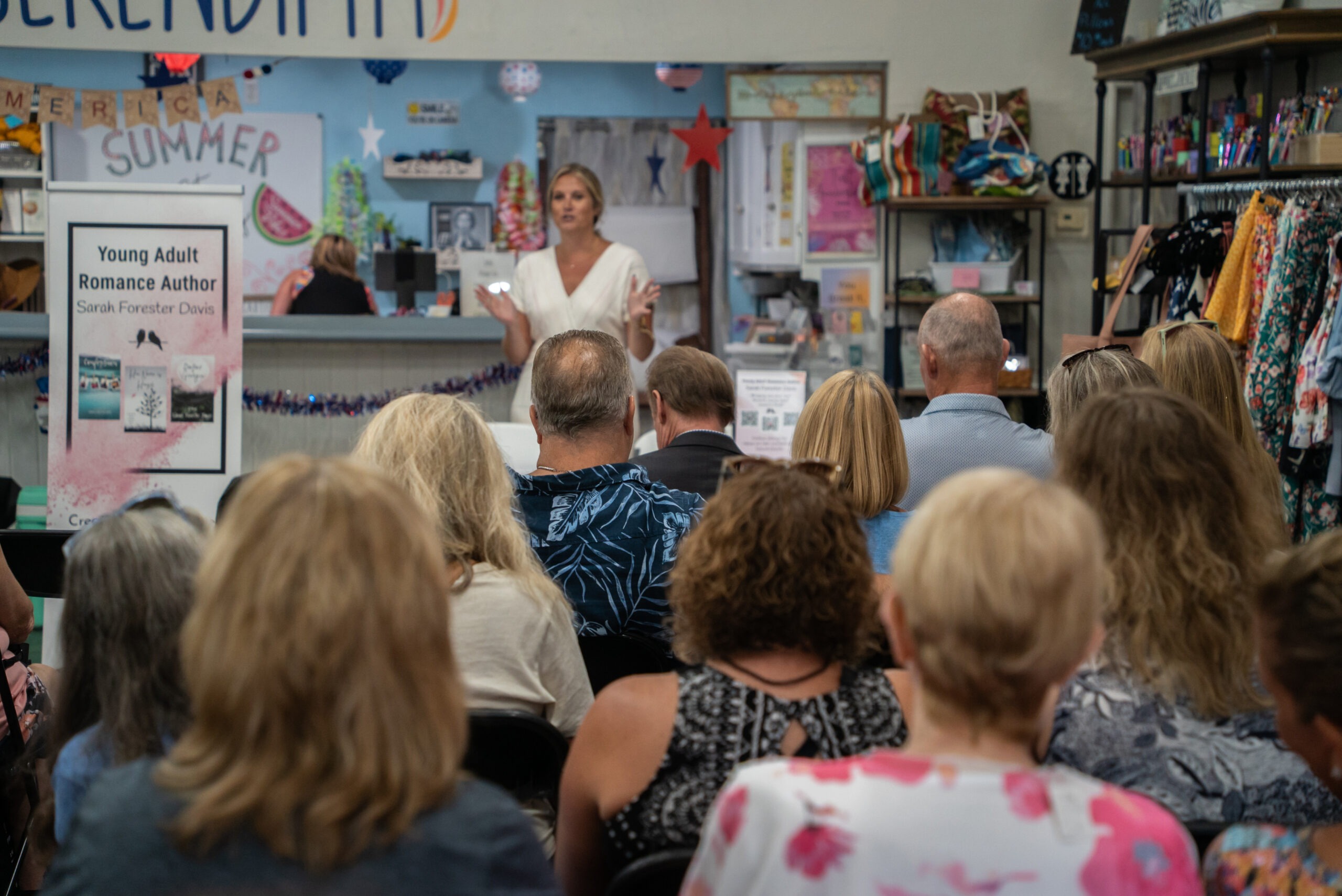 A woman stands at a podium in a bookstore, addressing an audience seated in front of her A sign next to her reads "Young Adult Romance Author Sarah Forster Davis " Shelves filled with books and colorful decorations surround the area