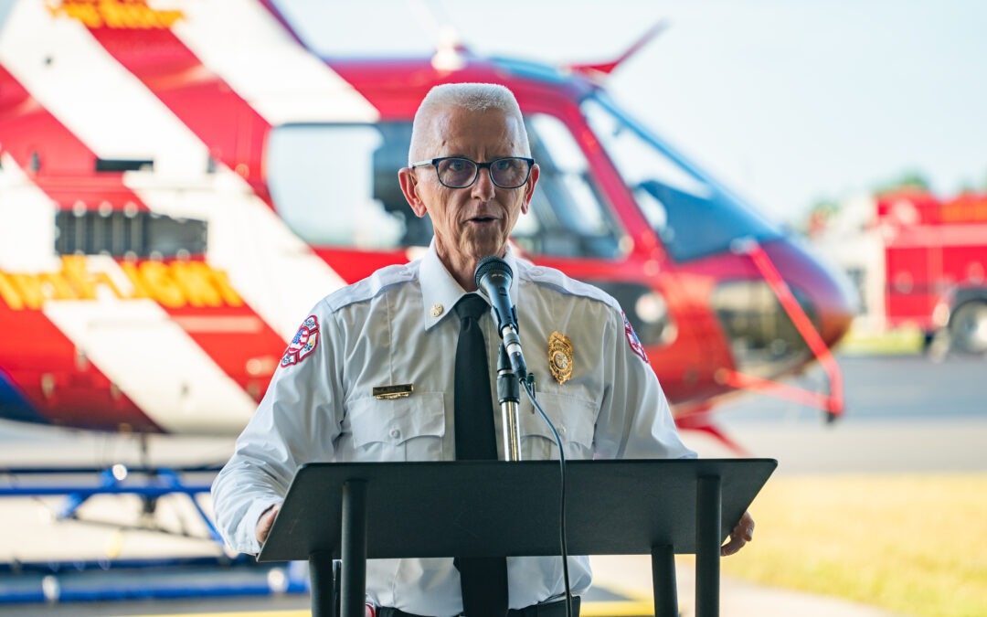 An elderly man with white hair and glasses, wearing a white uniform shirt with a fire department badge, is speaking at a podium Behind him is a red and white helicopter with emergency vehicles in the background